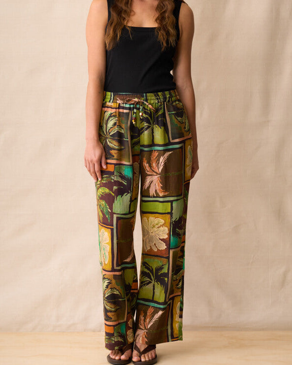 Neris model stands against a beige fabric backdrop wearing wide-leg pants with a colorful tropical patchwork print in green, brown, yellow, and orange, paired with a black sleeveless top and dark sandals.