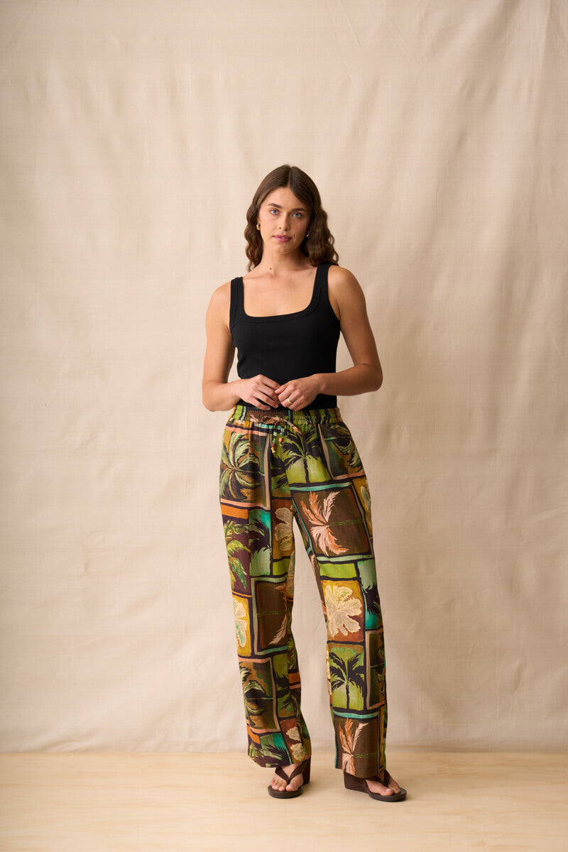 Neris model stands against a beige fabric backdrop wearing wide-leg pants with a colorful tropical patchwork print in green, brown, yellow, and orange, paired with a black sleeveless top and dark sandals.