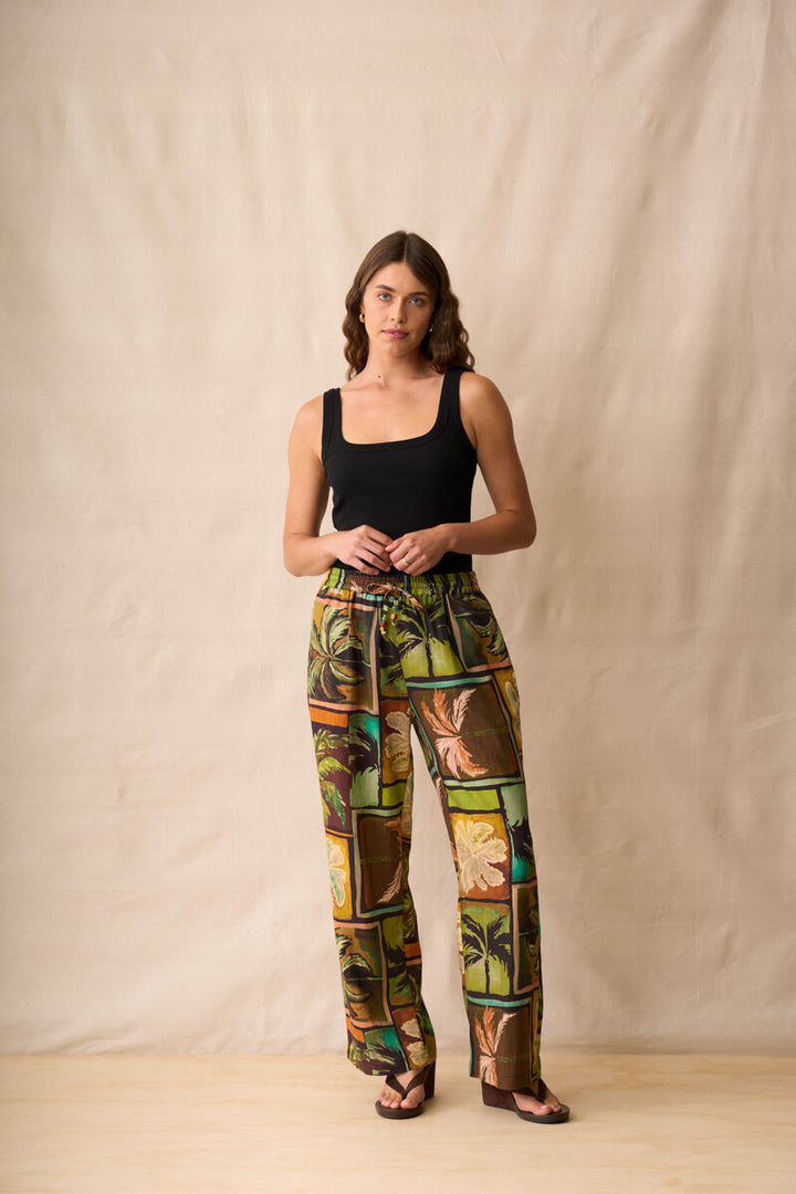 Neris model stands against a beige fabric backdrop wearing wide-leg pants with a colorful tropical patchwork print in green, brown, yellow, and orange, paired with a black sleeveless top and dark sandals.