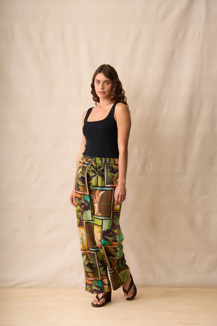 Neris model stands against a beige fabric backdrop wearing wide-leg pants with a colorful tropical patchwork print in green, brown, yellow, and orange, paired with a black sleeveless top and dark sandals.