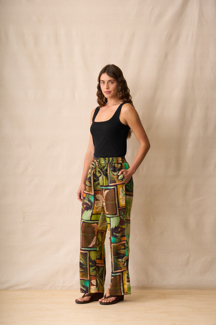 Neris model stands against a beige fabric backdrop wearing wide-leg pants with a colorful tropical patchwork print in green, brown, yellow, and orange, paired with a black sleeveless top and dark sandals.