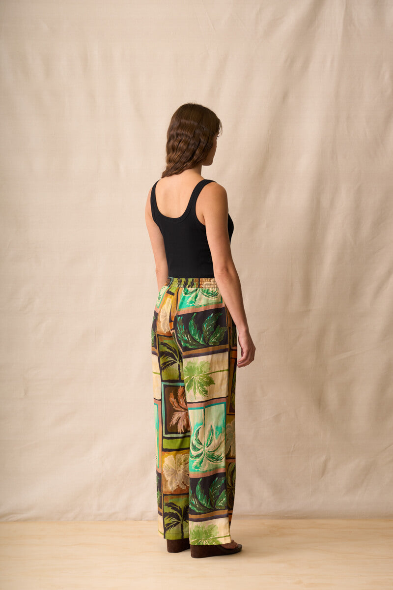 Neris model stands against a beige fabric backdrop wearing wide-leg pants with a colorful tropical patchwork print in green, brown, yellow, and orange, paired with a black sleeveless top and dark sandals. Back view