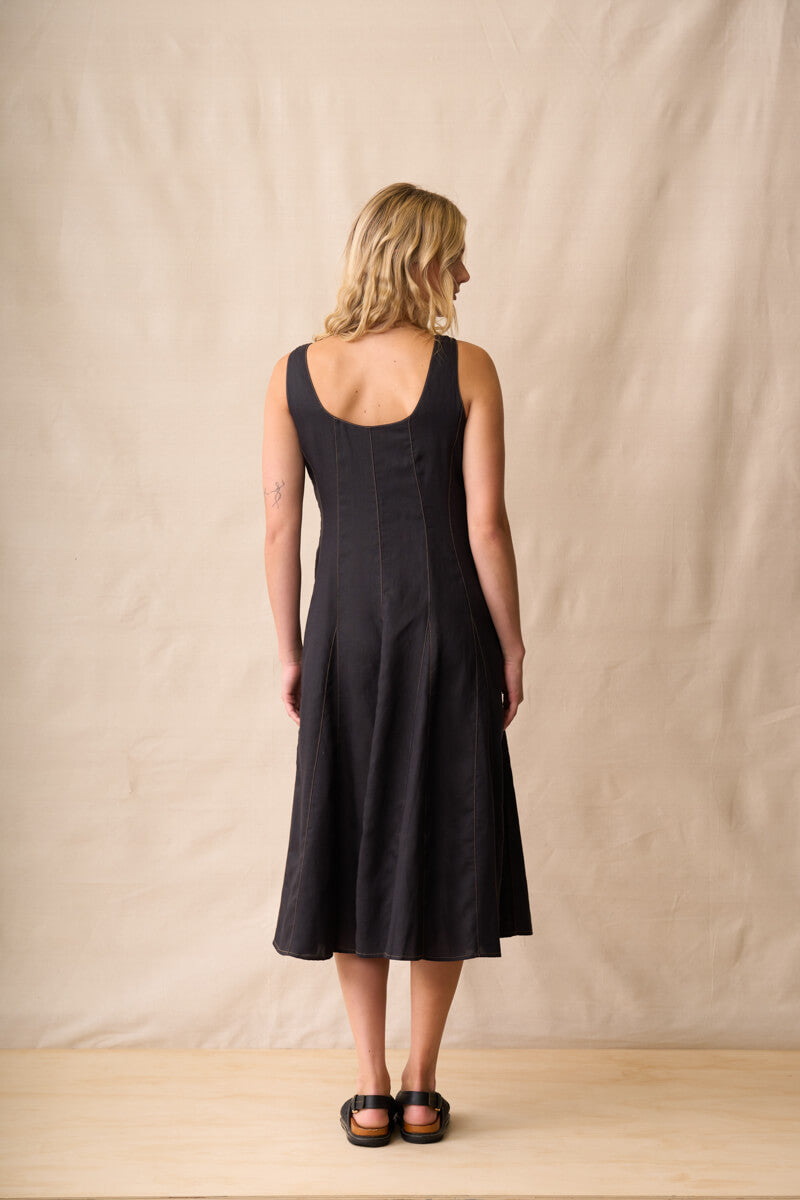 Neris modeling a black linen midi dress with a square neckline and wide shoulder straps, standing against a neutral beige fabric backdrop. Back view