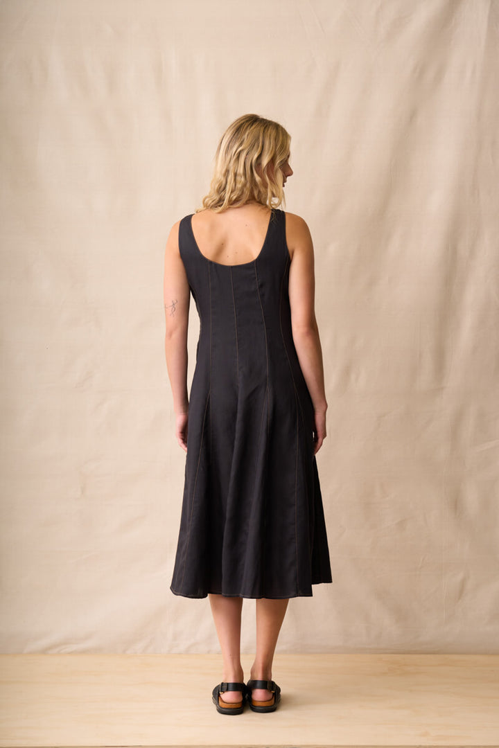 Neris modeling a black linen midi dress with a square neckline and wide shoulder straps, standing against a neutral beige fabric backdrop. Back view
