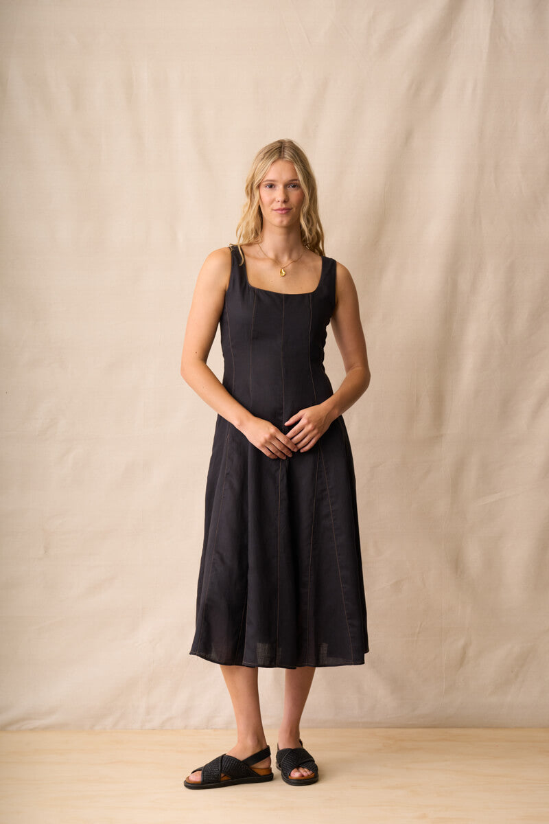 Neris modeling a black linen midi dress with a square neckline and wide shoulder straps, standing against a neutral beige fabric backdrop.