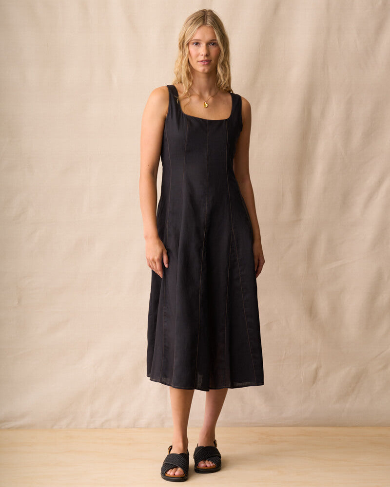 Neris modeling a black linen midi dress with a square neckline and wide shoulder straps, standing against a neutral beige fabric backdrop.