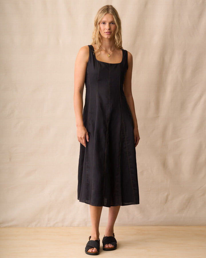 Neris modeling a black linen midi dress with a square neckline and wide shoulder straps, standing against a neutral beige fabric backdrop.