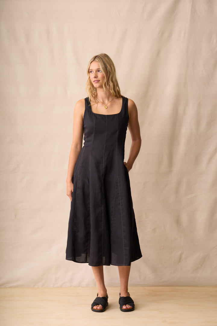 Neris modeling a black linen midi dress with a square neckline and wide shoulder straps, standing against a neutral beige fabric backdrop.
