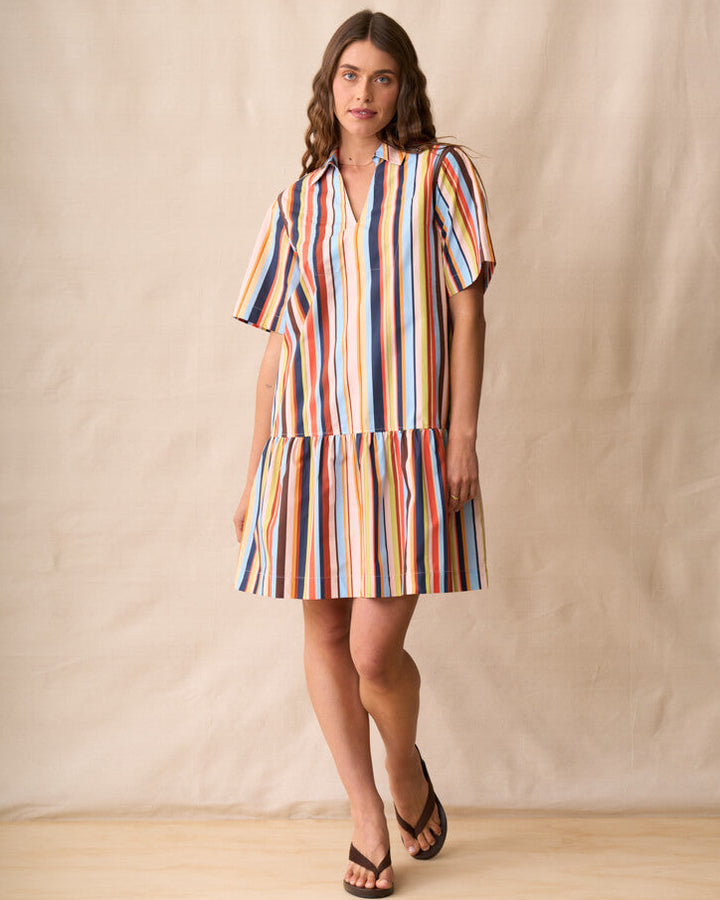 Neris modeling a short-sleeve, tiered mini dress with vertical multicolor stripes in red, orange, yellow, green, blue, and white, set against a neutral beige fabric backdrop.