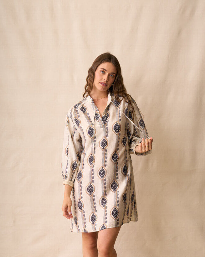 Neris modeling a long-sleeve button-up shirt dress with a beige, white, and navy geometric pattern, standing against a neutral beige fabric backdrop.
