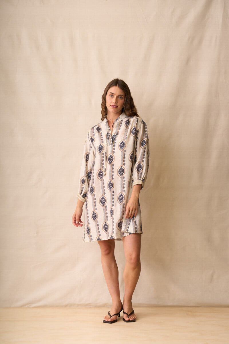 Neris modeling a long-sleeve button-up shirt dress with a beige, white, and navy geometric pattern, standing against a neutral beige fabric backdrop.