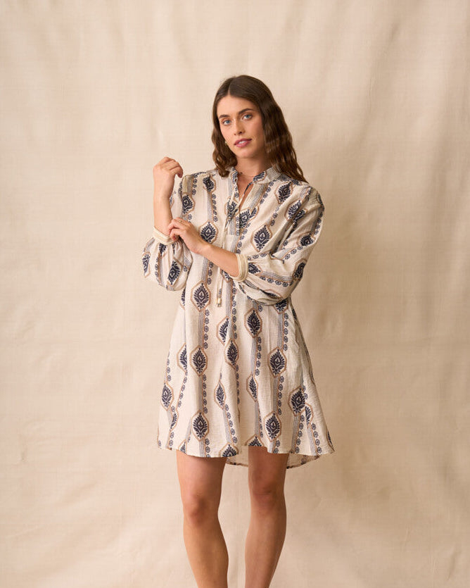 Neris modeling a long-sleeve button-up shirt dress with a beige, white, and navy geometric pattern, standing against a neutral beige fabric backdrop.