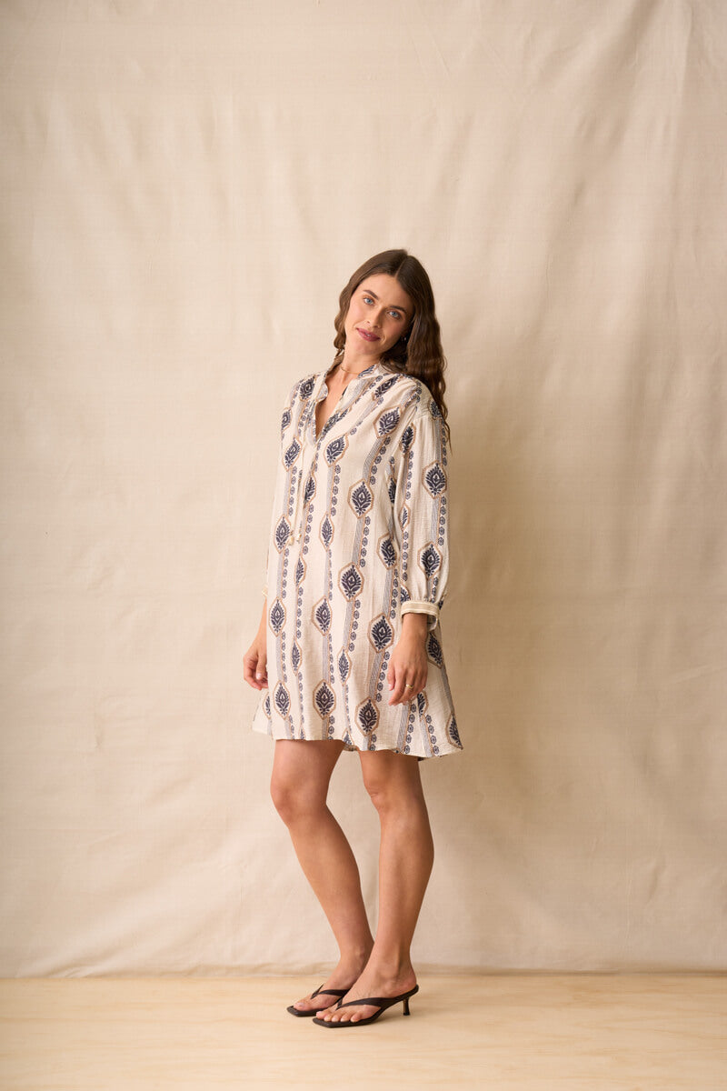 Neris modeling a long-sleeve button-up shirt dress with a beige, white, and navy geometric pattern, standing against a neutral beige fabric backdrop.