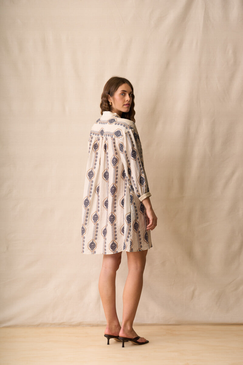 Neris modeling a long-sleeve button-up shirt dress with a beige, white, and navy geometric pattern, standing against a neutral beige fabric backdrop. Back view.