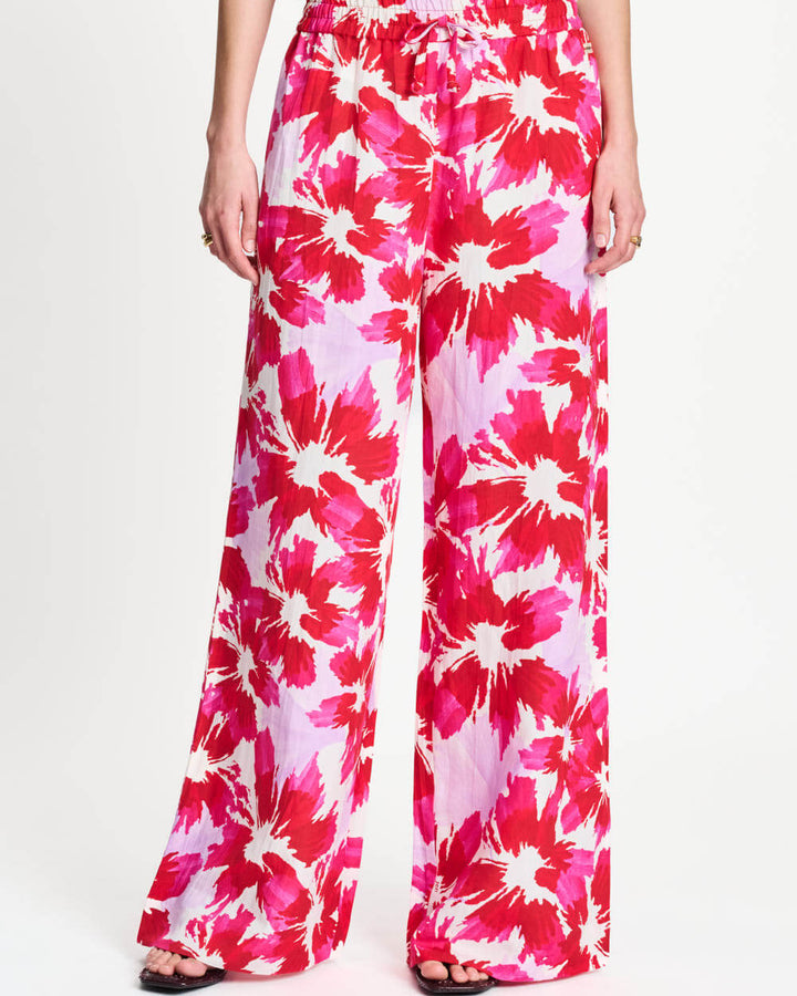 A model wearing high-waisted, wide-leg trousers with a vibrant pink, red, and white floral print, paired with a white graphic t-shirt and black sandals, against a plain white studio background.