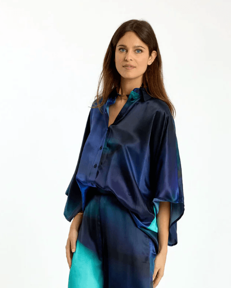 Lauren Vidal  modeling a matching blue and teal satin button-up shirt and trousers set against a plain white studio background.