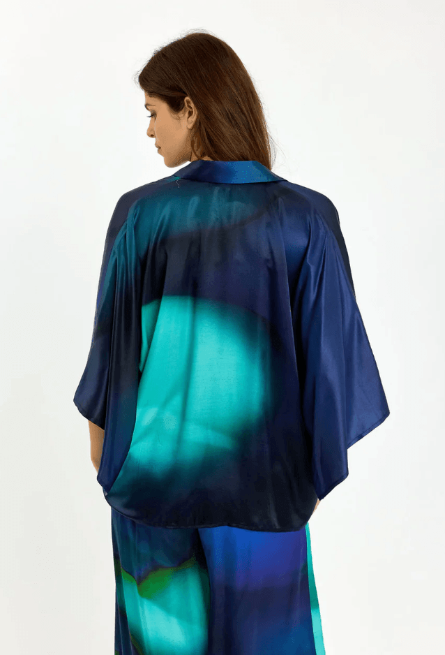 Lauren Vidal  modeling a matching blue and teal satin button-up shirt and trousers set against a plain white studio background. Back view.