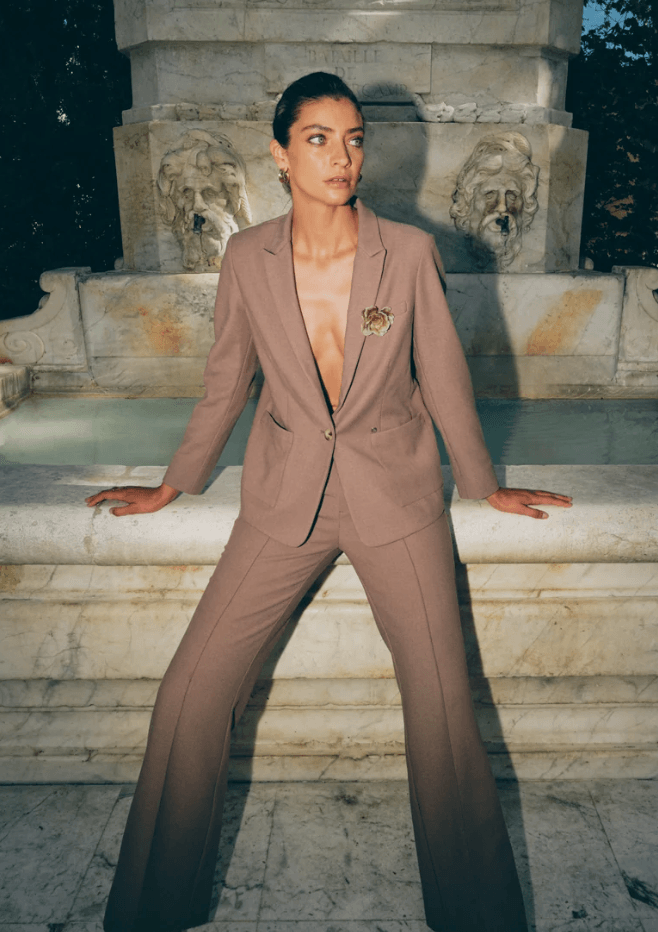 Mos Mosh  woman models a matching taupe brown two-piece suit with wide-leg trousers and a single-breasted blazer, posing in front of a stone fountain.