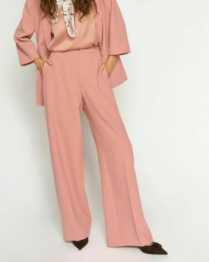 Mos Mosh A woman modeling a dusty pink two-piece suit consisting of a long-sleeve blazer and matching wide-leg trousers, styled with a patterned neck scarf and dark brown pointed-toe flats against a plain white studio background.
