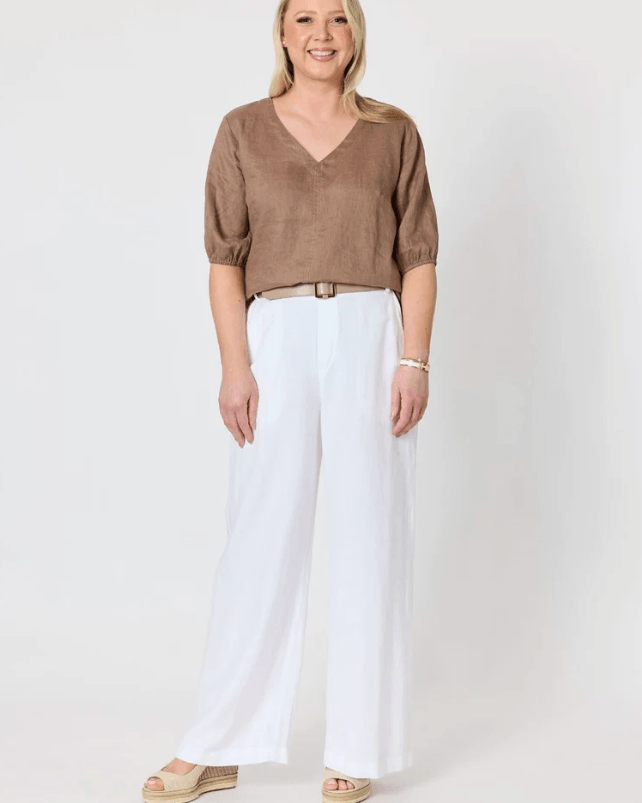 A woman modeling a brown short-sleeve V-neck top with white wide-leg pants and beige espadrille sandals, standing against a plain light gray studio background.