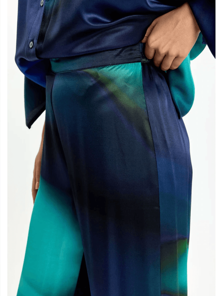 Lauren Vidal model wearing wide-leg trousers with an abstract teal, green, and navy blue gradient print, paired with a matching dark blue satin blouse and brown pointed-toe heels, shot against a plain white studio background. Close up.