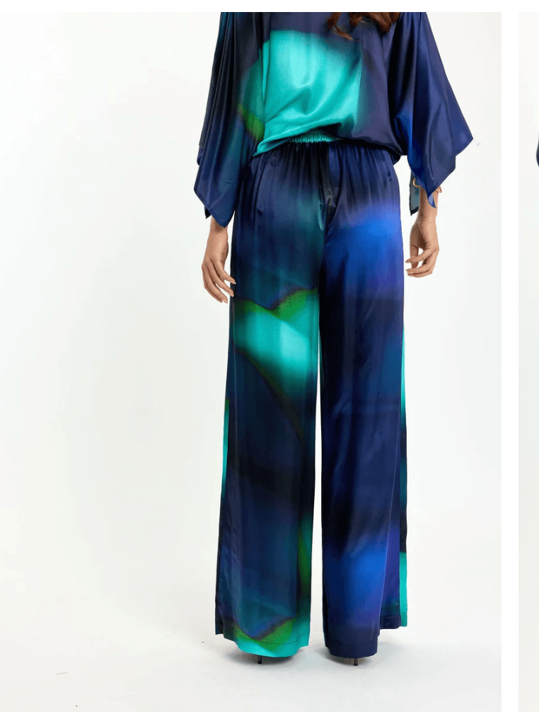 Lauren Vidal model wearing wide-leg trousers with an abstract teal, green, and navy blue gradient print, paired with a matching dark blue satin blouse and brown pointed-toe heels, shot against a plain white studio background. Back View 