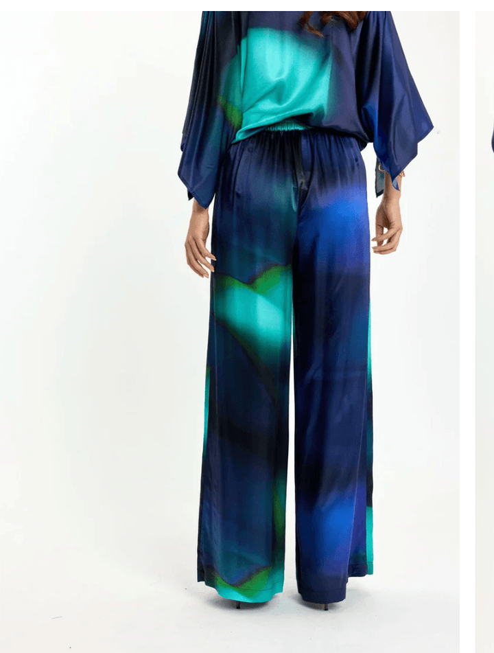 Lauren Vidal model wearing wide-leg trousers with an abstract teal, green, and navy blue gradient print, paired with a matching dark blue satin blouse and brown pointed-toe heels, shot against a plain white studio background. Back View 
