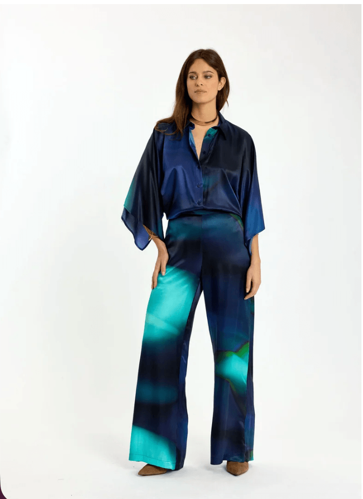 Lauren Vidal model wearing wide-leg trousers with an abstract teal, green, and navy blue gradient print, paired with a matching dark blue satin blouse and brown pointed-toe heels, shot against a plain white studio background.