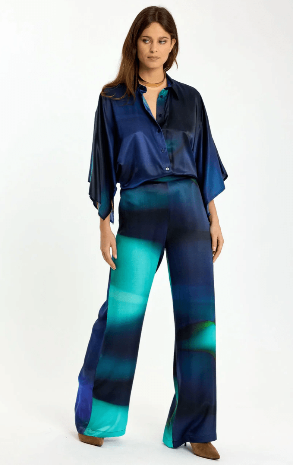 Lauren Vidal model wearing wide-leg trousers with an abstract teal, green, and navy blue gradient print, paired with a matching dark blue satin blouse and brown pointed-toe heels, shot against a plain white studio background.