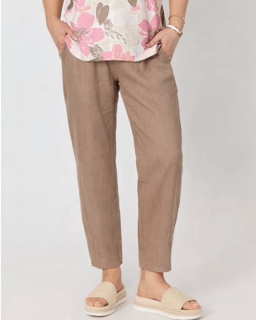 Gordon Smith model  wearing loose, straight-leg taupe brown trousers, paired with a floral top and beige slide sandals, standing against a light gray studio background.