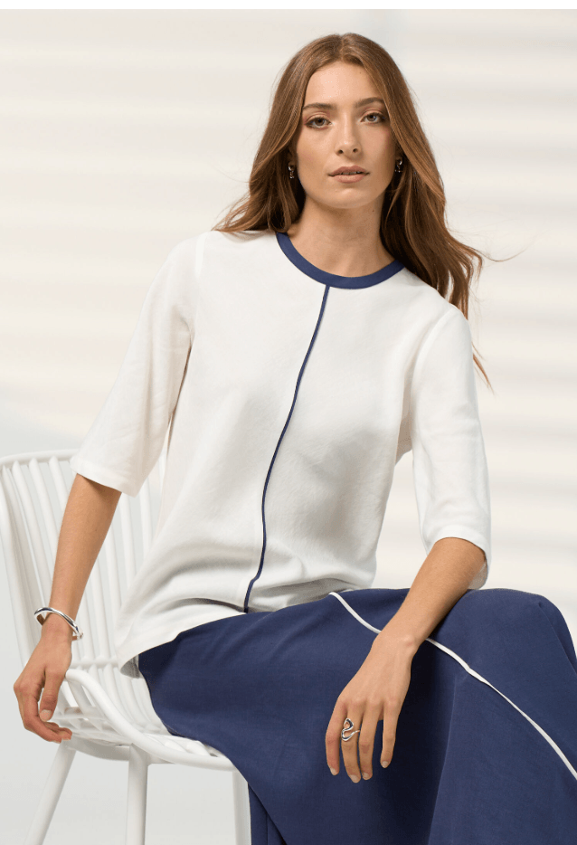 A woman seated on a white chair wearing a white short-sleeve top with navy trim and matching navy pants, photographed against a light neutral background.