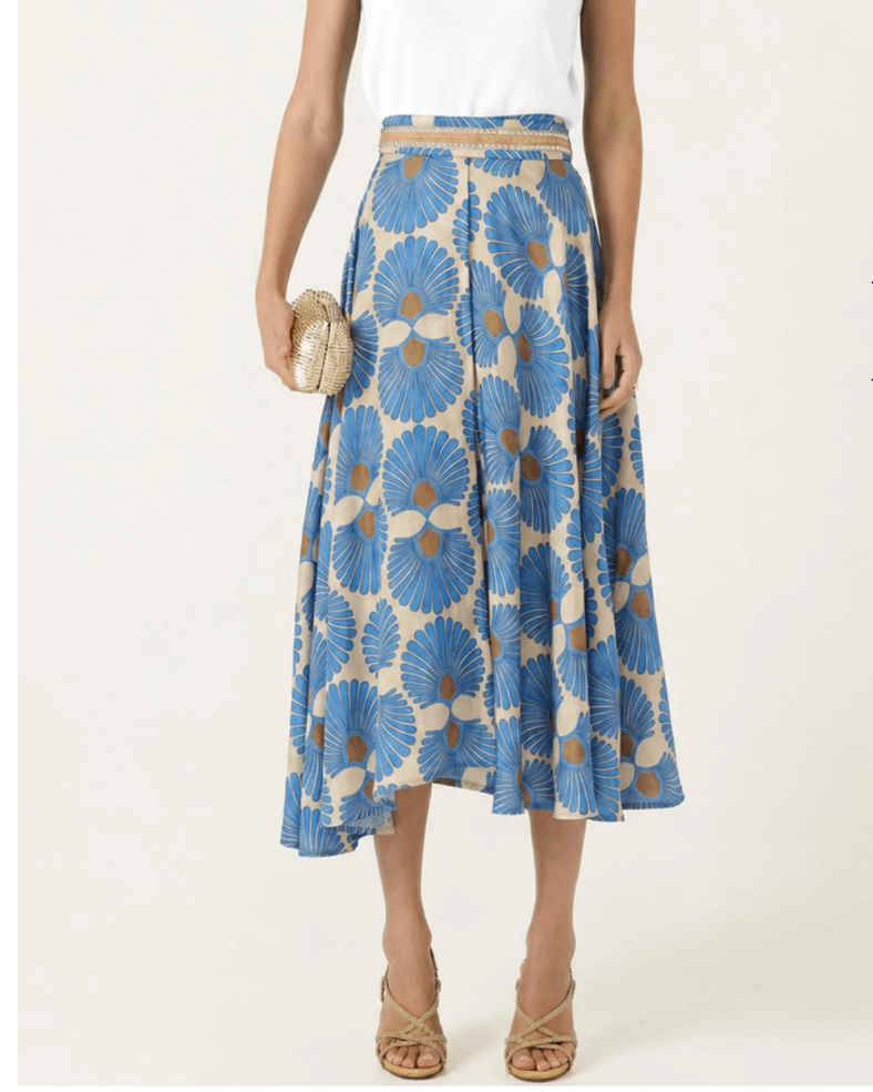 Sacha Drake  modeling a white sleeveless top with a textured chest panel and a high-waisted midi skirt featuring a blue, beige, and brown floral fan pattern, paired with beige strappy heels and holding a small metallic clutch. On a white studio background