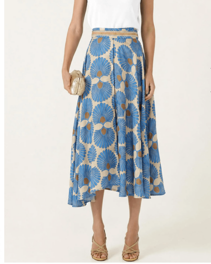 Sacha Drake  modeling a white sleeveless top with a textured chest panel and a high-waisted midi skirt featuring a blue, beige, and brown floral fan pattern, paired with beige strappy heels and holding a small metallic clutch. On a white studio background