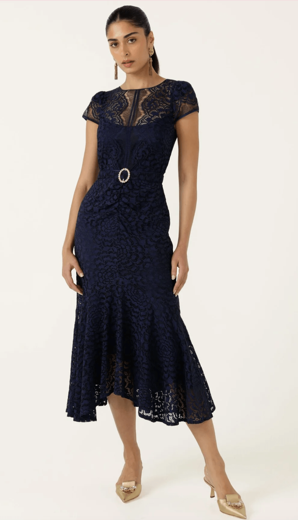 Sacha Drake models a navy blue lace midi dress with a round neckline, short cap sleeves, a matching belt with a silver ring, and a flared hem. She is standing outdoors against a beige stone wall.