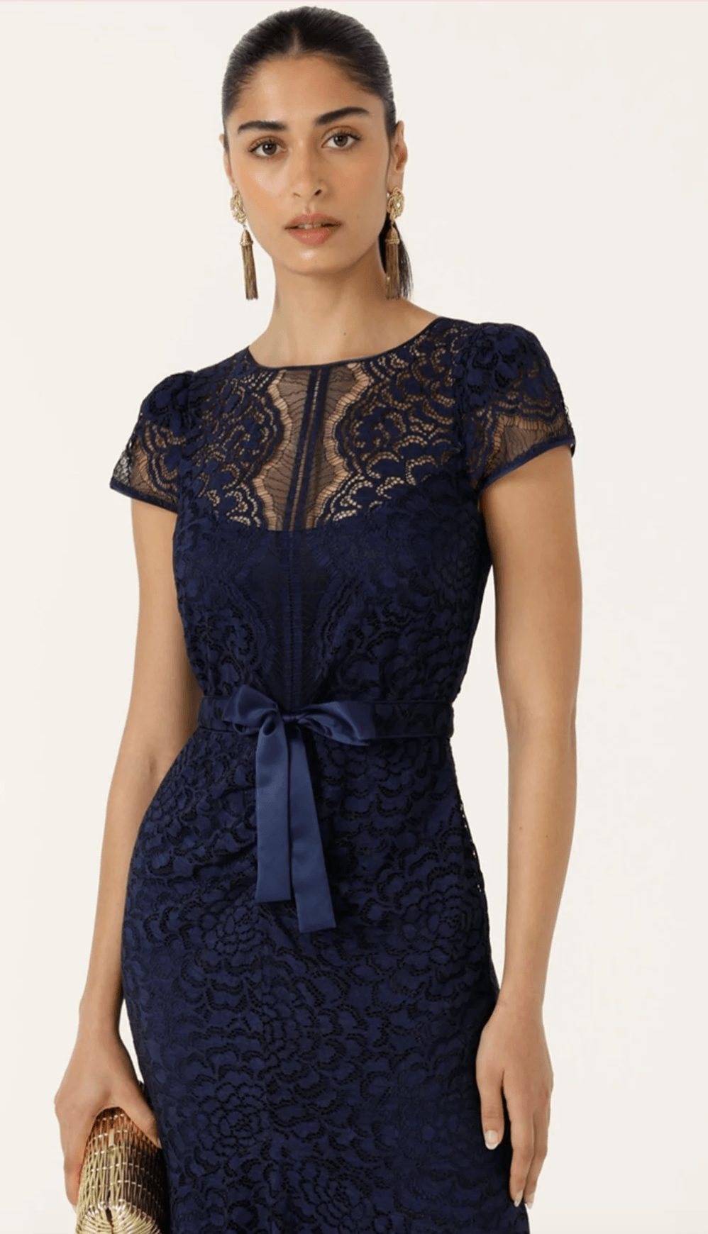 Sacha Drake models a navy blue lace midi dress with a round neckline, short cap sleeves, a matching belt with a silver ring, and a flared hem. She is standing outdoors against a beige stone wall.