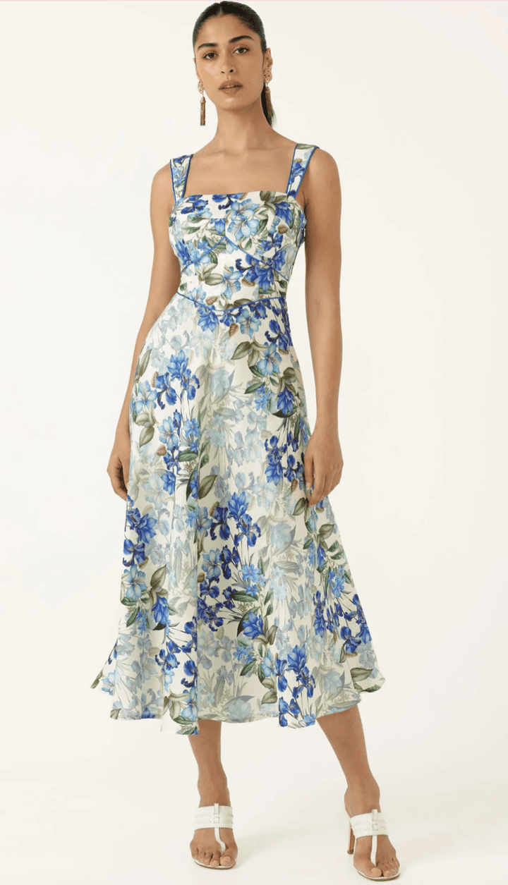 Sacha Drake  modeling a sleeveless midi dress with a blue and green floral print on a white background, paired with white flat sandals, against a plain white studio backdrop.