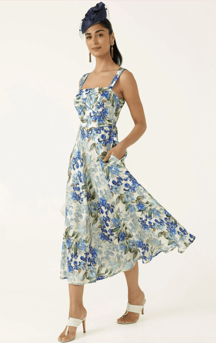 Sacha Drake  modeling a sleeveless midi dress with a blue and green floral print on a white background, paired with white flat sandals, against a plain white studio backdrop