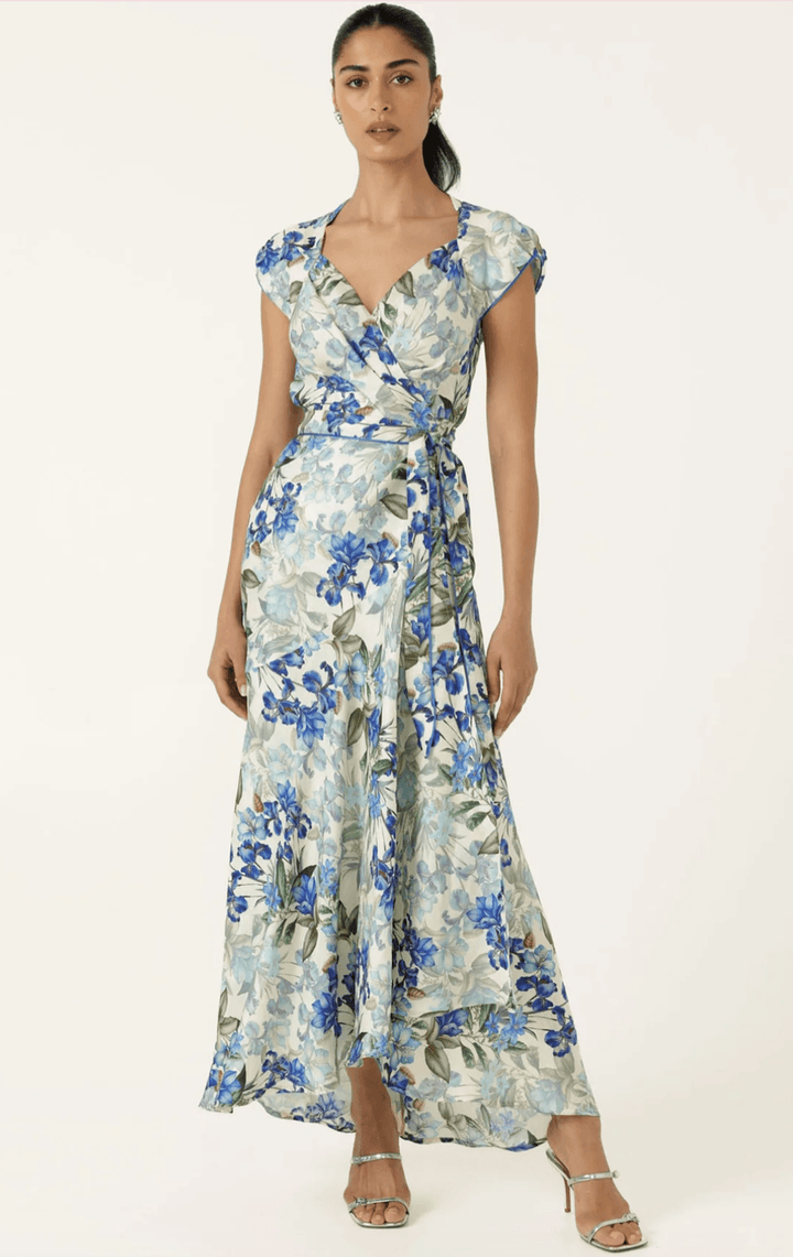 Sacha Drake modelling a white maxi dress with a blue and green floral print walks outdoors on a tan stone path with green grass and trees in the background. She is holding a small light blue clutch.