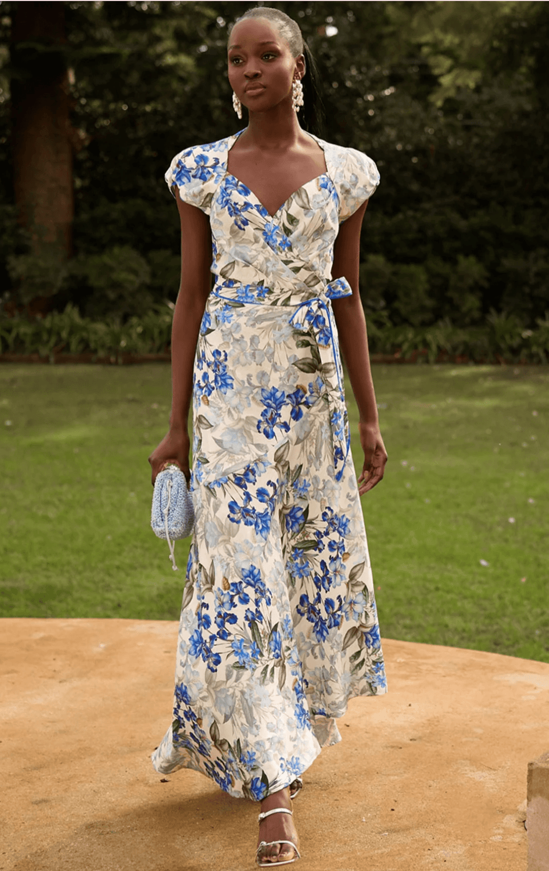 Sacha Drake modelling a white maxi dress with a blue and green floral print walks outdoors on a tan stone path with green grass and trees in the background. She is holding a small light blue clutch.