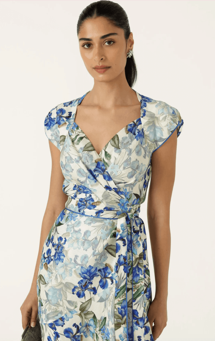 Sacha Drake modelling a white maxi dress with a blue and green floral print walks outdoors on a tan stone path with green grass and trees in the background. She is holding a small light blue clutch.