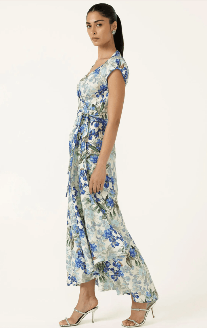 Sacha Drake modelling a white maxi dress with a blue and green floral print walks outdoors on a tan stone path with green grass and trees in the background. She is holding a small light blue clutch.