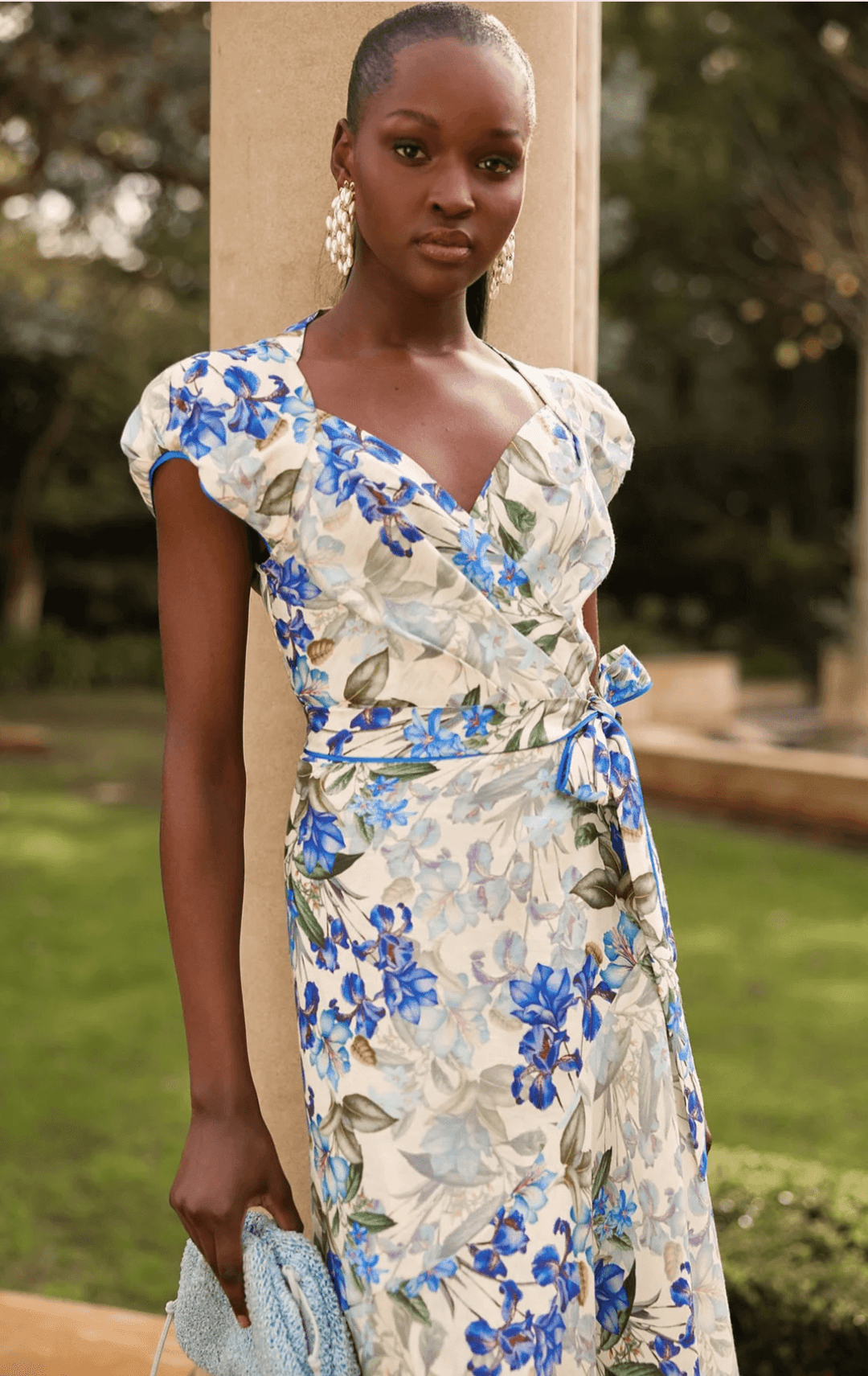 Sacha Drake modelling a white maxi dress with a blue and green floral print walks outdoors on a tan stone path with green grass and trees in the background. She is holding a small light blue clutch.