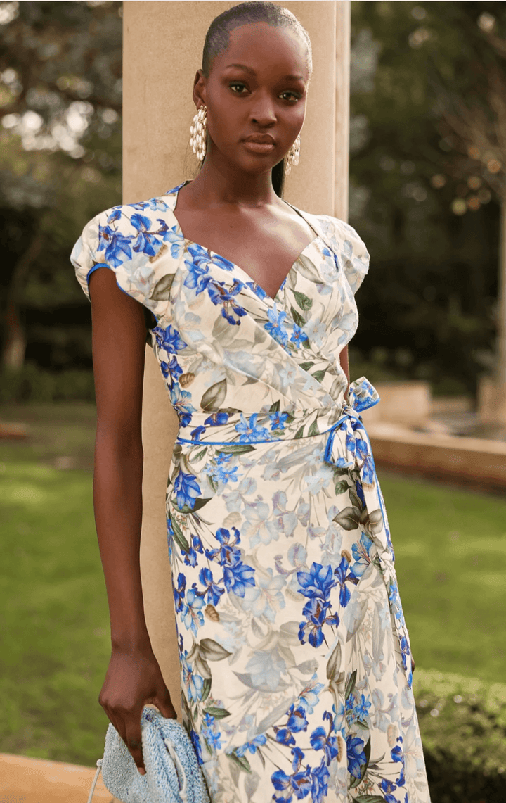 Sacha Drake modelling a white maxi dress with a blue and green floral print walks outdoors on a tan stone path with green grass and trees in the background. She is holding a small light blue clutch.