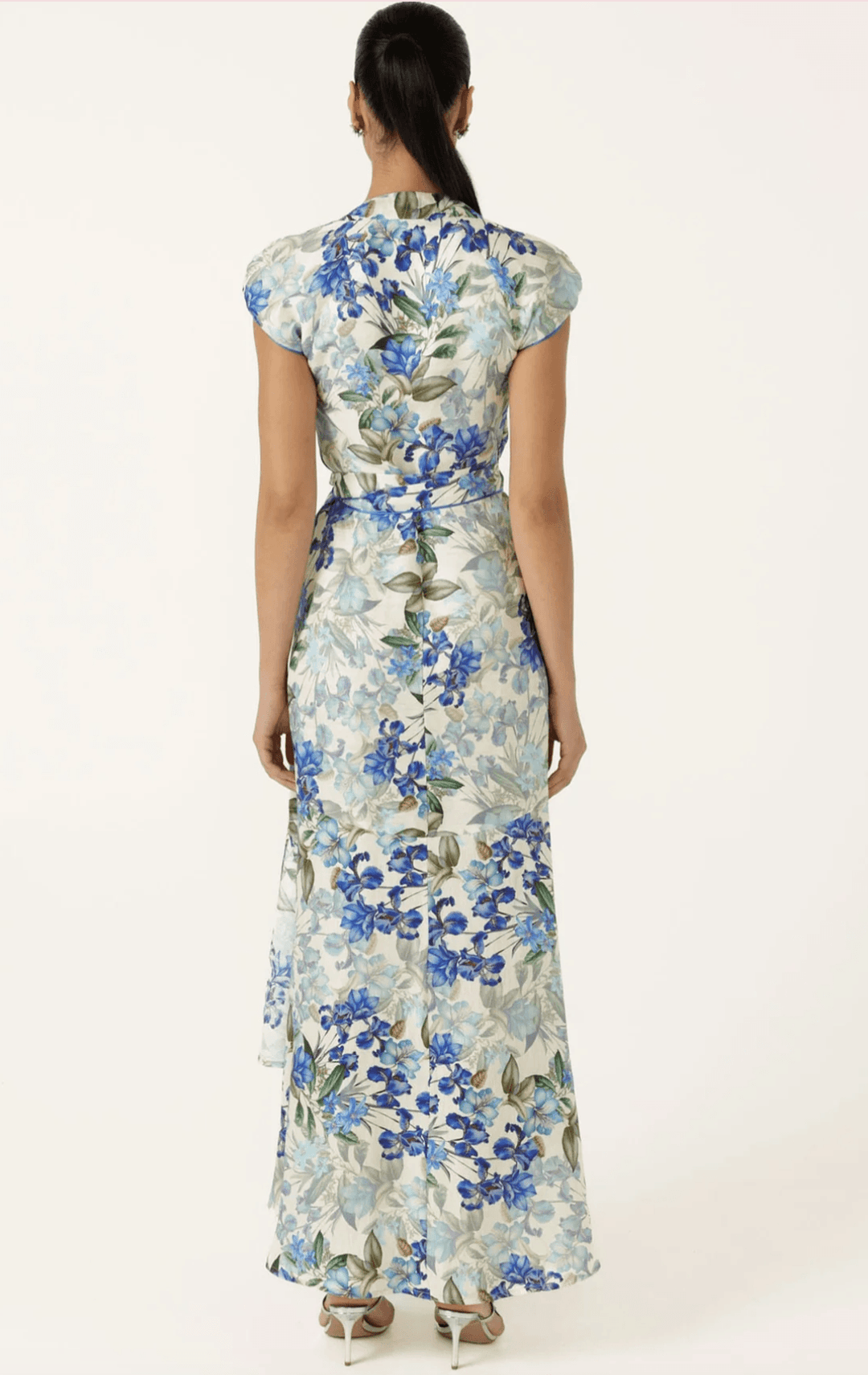 Sacha Drake modelling a white maxi dress with a blue and green floral print walks outdoors on a tan stone path with green grass and trees in the background. She is holding a small light blue clutch. Back view.
