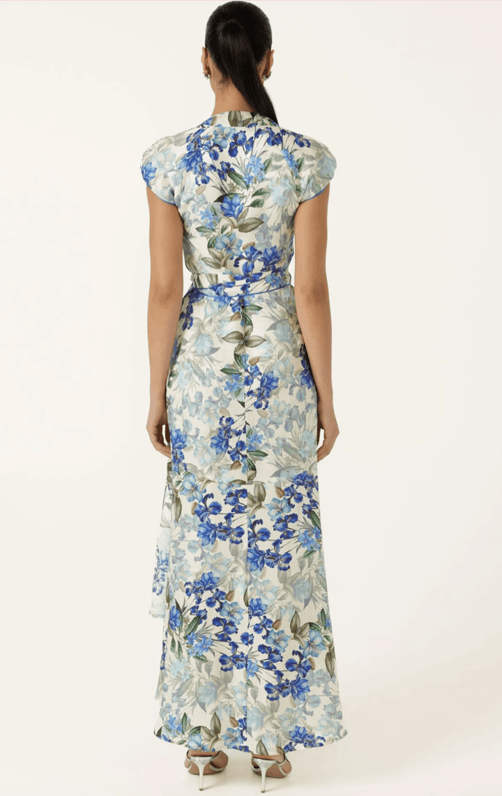 Sacha Drake modelling a white maxi dress with a blue and green floral print walks outdoors on a tan stone path with green grass and trees in the background. She is holding a small light blue clutch. Back view.