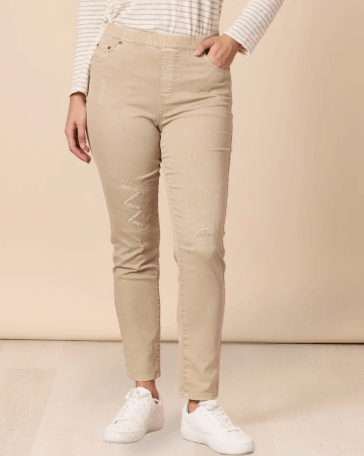 Threadz model  wearing beige slim-fit jeans with distressed details on the thighs, paired with a white and beige striped long-sleeve top and white sneakers, standing against a solid beige background.