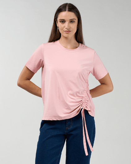 Madly Sweetly modeling a light pink short-sleeve t-shirt with a side ruching detail and tie, paired with dark blue jeans, against a light gray studio background.