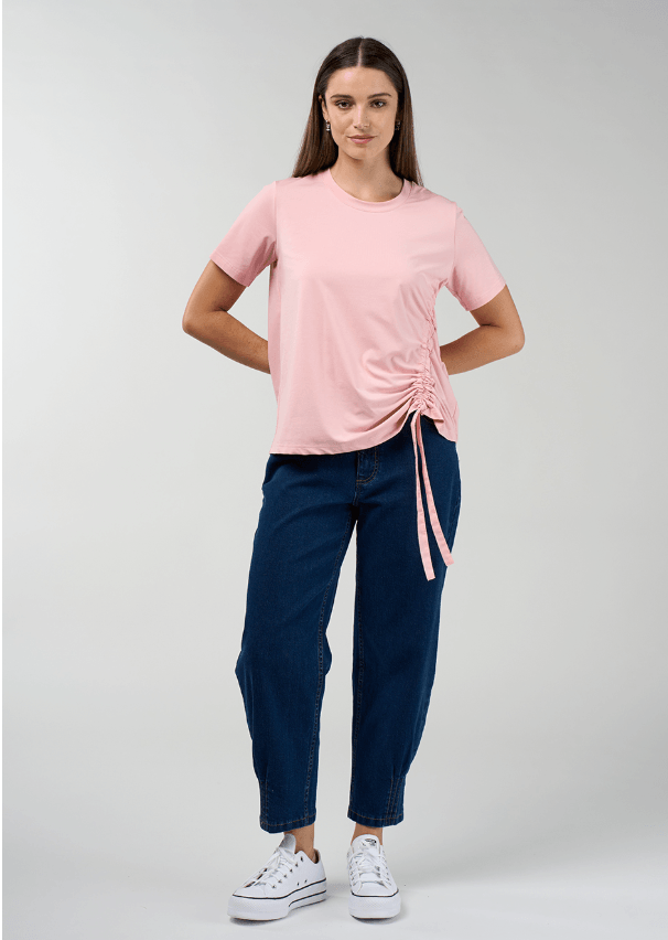 Madly Sweetly modeling a light pink short-sleeve t-shirt with a side ruching detail and tie, paired with dark blue jeans, against a light gray studio background.