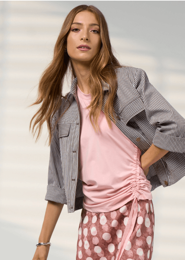 Madly Sweetly modeling a light pink short-sleeve t-shirt with a side ruching detail and tie, paired with dark blue jeans, against a light gray studio background.