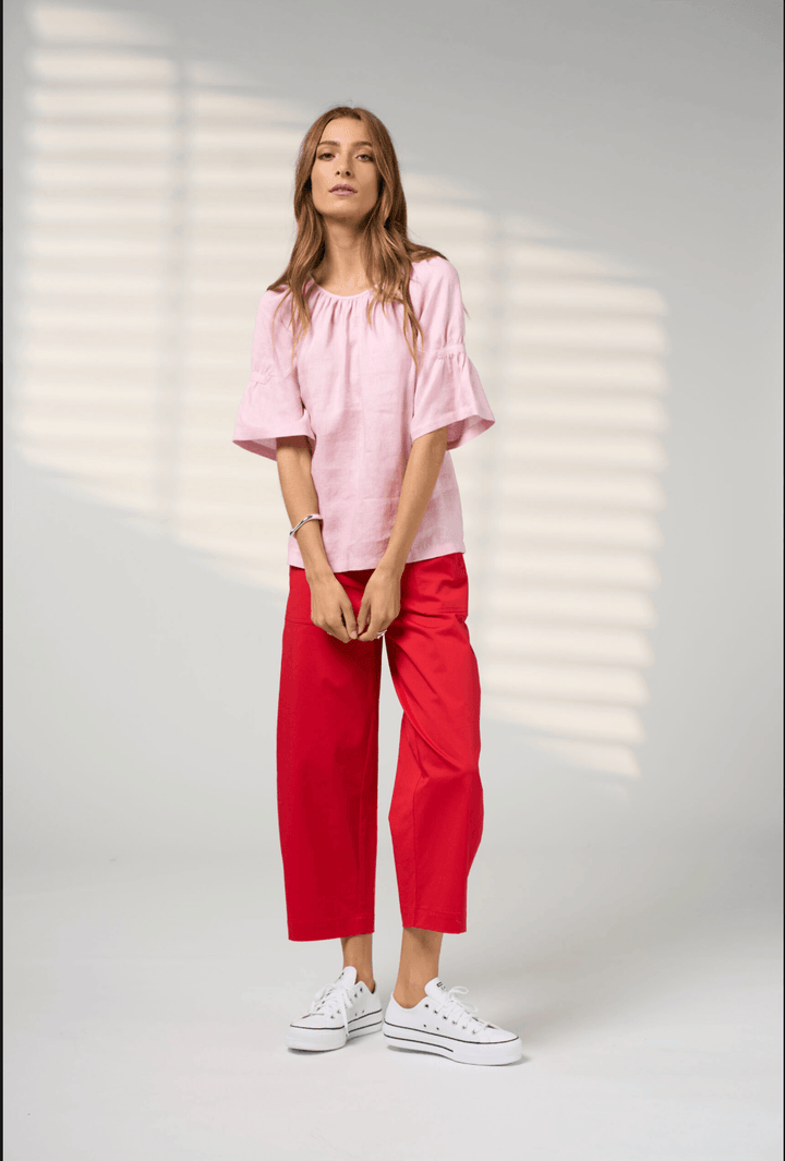 Madly Sweetly model wearing a light pink linen-blend top with a gathered round neckline and flared elbow-length sleeves, paired with blue jeans, set against a soft gray background with subtle leaf shadows.
